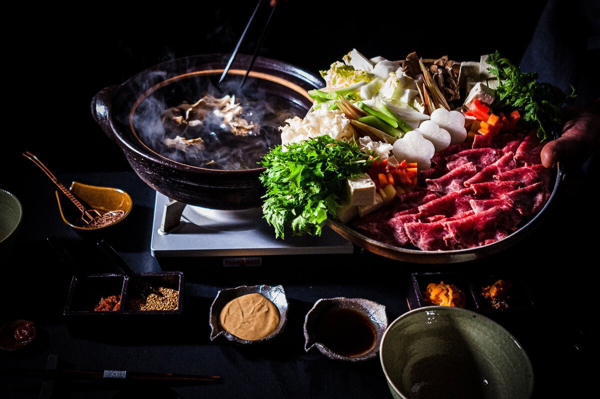 A steaming hot pot of broth surrounded by thinly sliced beef and farm vegetables. Empty bowls for serving, small dishes of sauce and other sides surround the large hot pot.